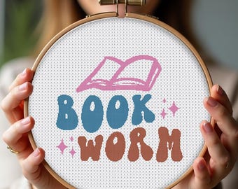 May include: A white embroidery hoop with a pink book illustration and the text "BOOK WORM" in blue and brown letters with a retro font. The letters are outlined in white.