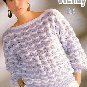 May include: A light blue and white knitted sweater with a lacy pattern. The sweater has a boat neck and long sleeves. The sweater is shown on a model wearing white pants.