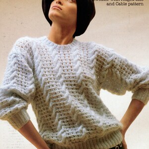 May include: A light blue knitted sweater with a cable and lace pattern. The sweater has a dropped shoulder line and is shown on a model wearing a black turban. The text "Romance" is printed in pink at the top of the image. The text "C 6985 30-40" is printed in black at the top right of the image. The text "Dropped shoulder line Sweater with ridged lace and Cable pattern" is printed in black at the right side of the image.
