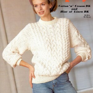 May include: A white knitted sweater with a cable knit pattern. The sweater is worn by a model with a colorful scarf and blue jeans. The text on the image reads "Cotton'n' Cream DK and Hint of Linen DK 30-44 in 76-112 cm".