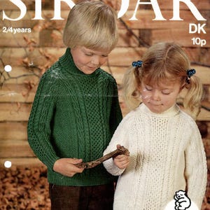 May include: Vintage Sirdar knitting pattern cover featuring two children in knitted sweaters. The boy wears a green cable-knit sweater, and the girl wears a white sweater. The text reads "Sirdar" and "Snuggly Baby Yarns".