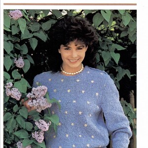 May include: A light blue sweater with small yellow flower-shaped accents. The image also features the text "Lilac time" and "Quick and Easy Fashions". The sweater is worn with white pants and a pearl necklace.
