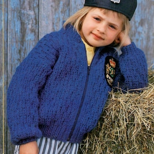 May include: A blue knitted cardigan with a zip fastening and a decorative patch on the front. The cardigan is being worn by a child wearing a black baseball cap with a crest on it. The child is standing next to a bale of hay.