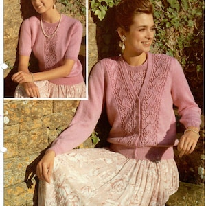 May include: A pink knitted cardigan with a V-neck and a button closure. The cardigan has a delicate cable knit pattern and is worn over a matching pink knitted top. The model is wearing a floral skirt and is standing against a brick wall with ivy.