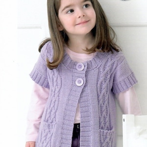Puede incluir: Una niña pequeña lleva un cárdigan de punto lavanda con un patrón de punto de cable y dos botones. El cárdigan tiene mangas cortas y bolsillos. La niña lleva una camisa rosa y pantalones morados.