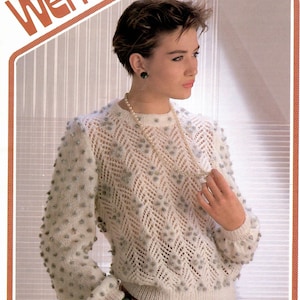 Puede incluir: Un suéter de punto blanco con un patrón de encaje y pequeños pompones blancos. El suéter tiene un cuello redondo y mangas largas. La modelo lleva un collar de perlas y una pulsera verde.