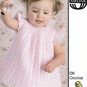 May include: A pink crocheted baby dress with a scalloped hem. The dress is made with a delicate stitch pattern and has short sleeves. The dress is for a baby girl aged 19-22 inches. The pattern is from Sirdar, a brand of yarn.