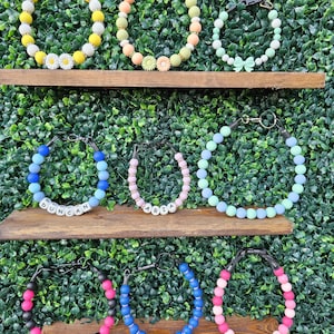 May include: A collection of colorful beaded pet collars displayed on wooden shelves against a green hedge background. The collars feature various colors, including yellow, pink, blue, and green, with some adorned with daisy and bow accents. Some collars have names spelled out.