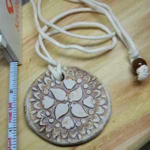 May include: A white ceramic pendant with a brown floral design hangs from a white cord with a brown wooden bead at the end.