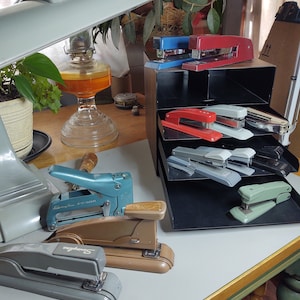 Puede incluir: Una colección de grapadoras vintage en varios colores, incluyendo azul, rojo, gris y verde. Algunas grapadoras se exhiben en una estantería escalonada, mientras que otras están sobre una superficie blanca. La imagen también incluye una lámpara de escritorio y una lámpara de aceite de vidrio.