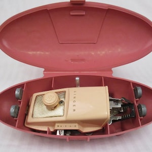 May include: A vintage Singer sewing machine in a pink carry case. The machine is beige with a gold-coloured dial and the Singer logo. The case is open, revealing the machine and its components. The case has wheels.