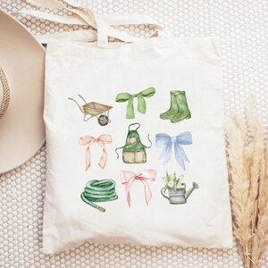 May include: Cream-colored tote bag featuring watercolor illustrations of gardening tools and accessories. The design includes a wheelbarrow, bows, boots, an apron, a hose, and a watering can. The illustrations are in shades of green, pink, and blue.