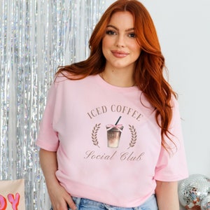 May include: A light pink t-shirt featuring the text "ICED COFFEE Social Club" in brown script. The design includes a watercolor illustration of an iced coffee drink with a pink bow and a black straw, surrounded by a laurel wreath.