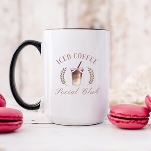 May include: White ceramic mug with a black handle and rim, featuring the text "Iced Coffee Social Club" in brown script. The design includes an illustration of an iced coffee drink with a pink bow and a straw.