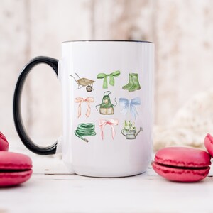 May include: White ceramic mug with a black handle and rim, featuring watercolor illustrations of gardening tools and bows. The design includes a wheelbarrow, boots, apron, and watering can. The mug is surrounded by pink macarons.