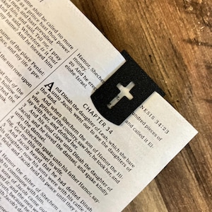 May include: A black bookmark with a cross cut out in the centre. The bookmark is on a page of a Bible open to Chapter 34 of Genesis. The text on the page reads "And Dinah the daughter of Leah, which she bare unto Jacob, went out to see the daughters of the land."