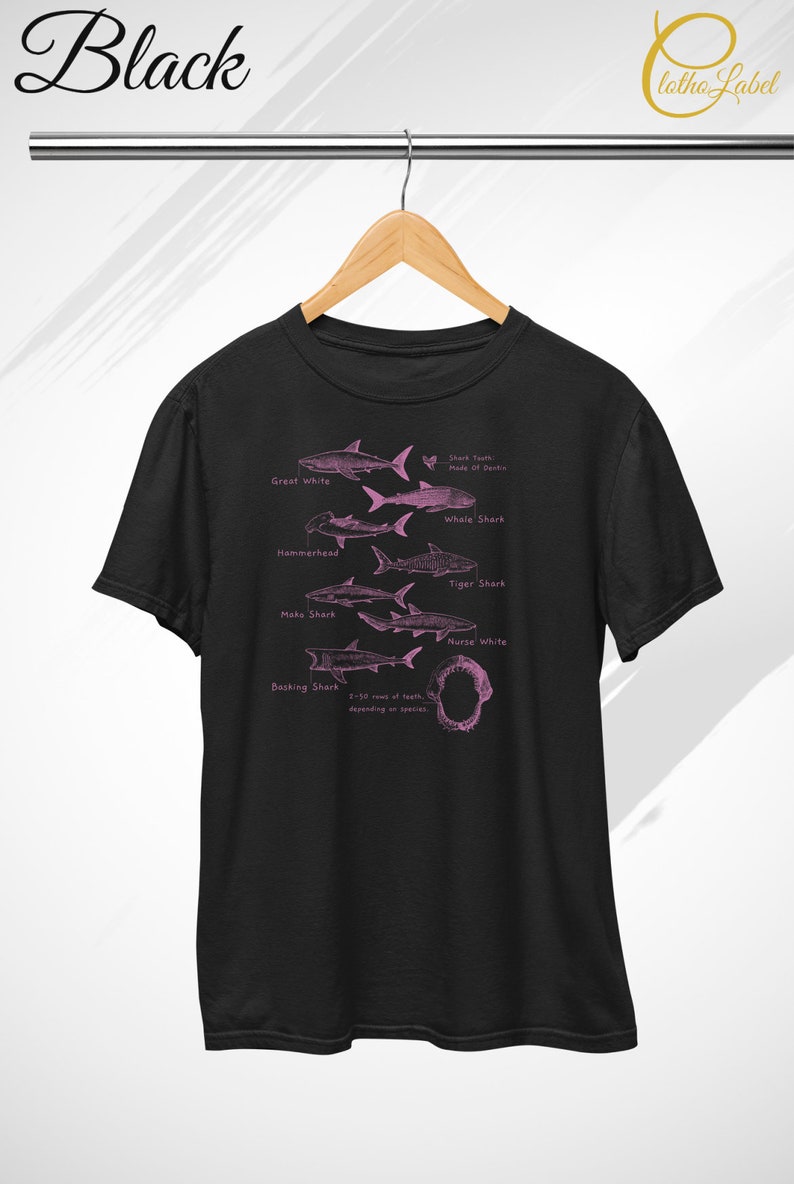 Pode incluir: Camiseta preta com uma ilustra&ccedil;&atilde;o rosa de diferentes esp&eacute;cies de tubar&otilde;es, incluindo o tubar&atilde;o branco, o tubar&atilde;o baleia, o tubar&atilde;o martelo, o tubar&atilde;o tigre, o tubar&atilde;o mako, o tubar&atilde;o-enfermeira e o tubar&atilde;o-peregrino. A ilustra&ccedil;&atilde;o inclui o texto "Shark Teeth Made of Dentine" e "2-10 rows of teeth, depending on species".