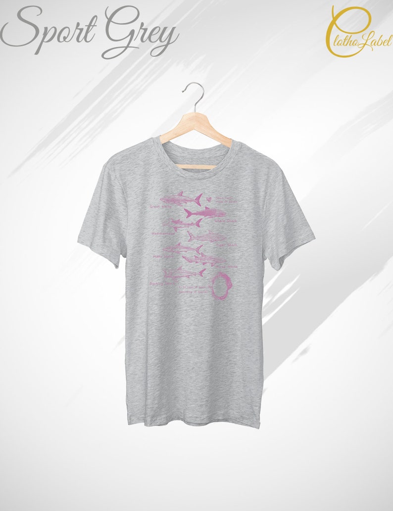 Pode incluir: Uma camiseta cinza mesclada com um gr&aacute;fico rosa de diferentes tipos de tubar&otilde;es. O texto "Grande branco", "Mako", "Tigre", "Touro", "Martelo" e "Tubar&atilde;o baleia" est&aacute; impresso abaixo dos tubar&otilde;es.