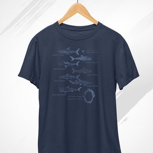 May include: A navy blue t-shirt with a graphic of different shark species, including the Great White, Whale Shark, Hammerhead, Tiger Shark, Mako Shark, Nurse Shark, and Basking Shark. The graphic also includes a shark tooth with the text "Shark Teeth Made of Dentine".
