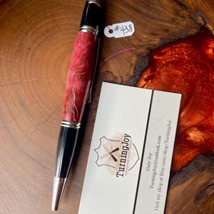 May include: A handcrafted pen with a red and gold patterned barrel, black accents, and silver-colored metal trim. The pen is next to a business card with the TurningJoy logo and contact information. A tag with the number #438 is attached.