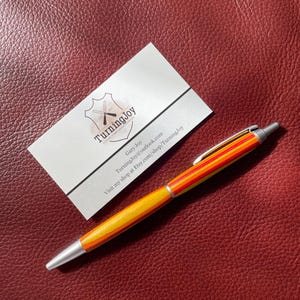 以下が含まれることがあります： 黄色、オレンジ、赤のストライプ模様のペン。先端とクリップはシルバーです。「TurningJoy」の文字と連絡先が書かれた名刺がペンの隣にあります。背景は、質感のある赤茶色の表面です。
