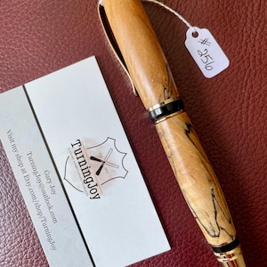May include: A hand-turned wooden pen with a natural light brown grain and dark streaks, featuring gold-tone metal accents, a black tip, and a white tag labeled #256. Beside it is a business card with the text Turningjoy, Gary Joy, and shop details.