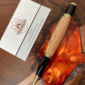 May include: A handcrafted wooden pen with a light brown barrel and black accents. The pen features gold-colored metal trim and a gold-colored tip. A business card with the TurningJoy logo is next to the pen.