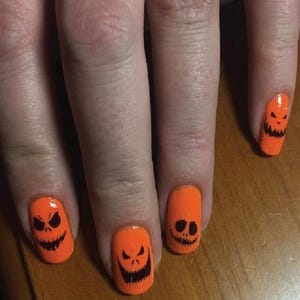 May include: Orange nail polish with black jack-o'-lantern faces painted on each nail. The faces have different expressions, including angry and smiling. The nails are on a hand.
