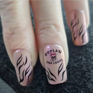 May include: Close-up of painted fingernails. The nails are a light pink with black flame designs. One nail features the text "OUTLAW JEEP LADIES" with a skull and bow graphic.