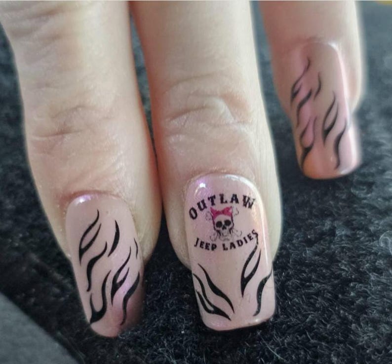 May include: Close-up of painted fingernails. The nails are a light pink with black flame designs. One nail features the text "OUTLAW JEEP LADIES" with a skull and bow graphic.