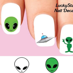 May include: Nail decals featuring cartoon aliens and a UFO on a white background. The aliens are green with black eyes and smiling faces. One alien is giving a peace sign. The UFO is silver and blue. The text "LuckyStar Nail Decals" is in the upper right corner.