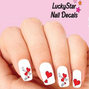 May include: White nail decals featuring red heart designs with black swirling flourishes. The text LuckyStar Nail Decals is printed in black above the nails, accompanied by three small stars in pink and orange colours on a light pink background.
