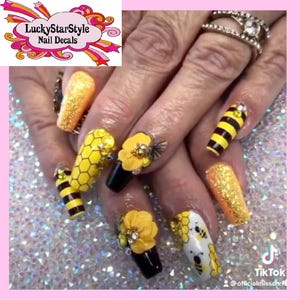 May include: A close-up of a hand with elaborately decorated nails. The nails feature a bee theme, with yellow and black stripes, honeycomb patterns, and 3D flower and bee embellishments. Some nails are covered in gold glitter. The LuckyStarStyle Nail Decals logo is visible.