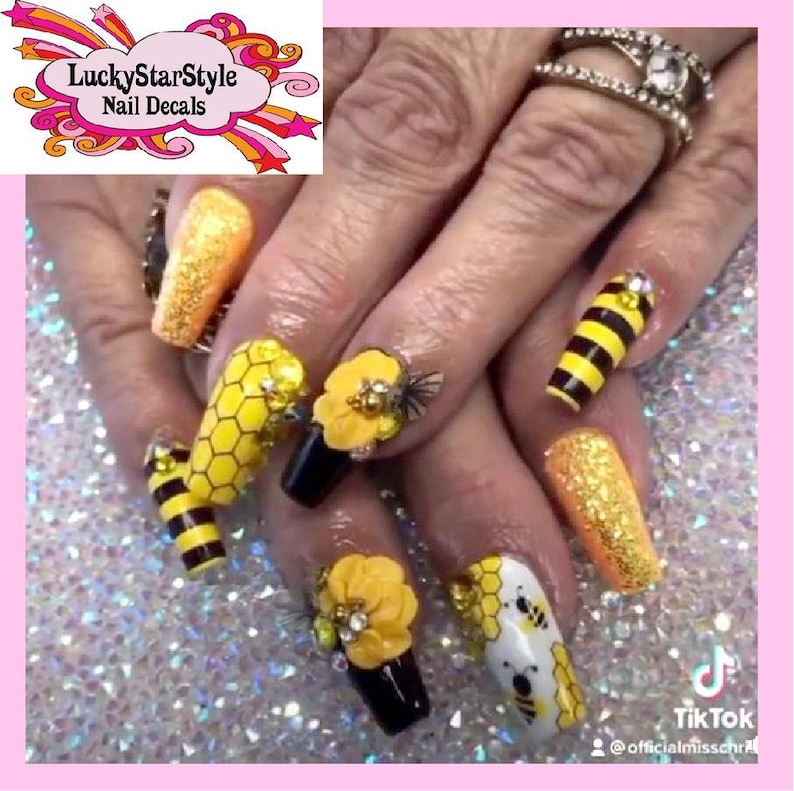 May include: A close-up of a hand with elaborately decorated nails. The nails feature a bee theme, with yellow and black stripes, honeycomb patterns, and 3D flower and bee embellishments. Some nails are covered in gold glitter. The LuckyStarStyle Nail Decals logo is visible.