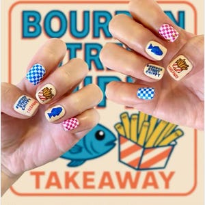 May include: A close-up of hands with nail art featuring a takeaway theme. The nails display images of fish, fries, and checkered patterns in blue, pink, and white. The background includes the words "BOURDOR STREET CHIPPY TAKEAWAY" and a blue fish illustration.