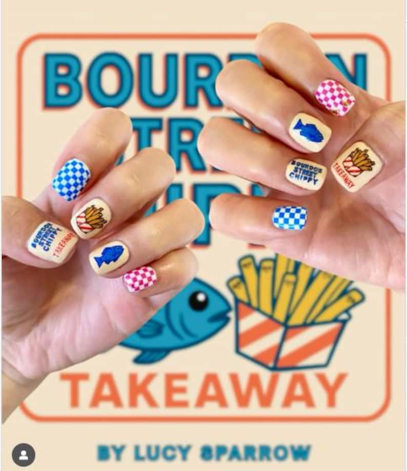 May include: A close-up of hands with nail art featuring a takeaway theme. The nails display images of fish, fries, and checkered patterns in blue, pink, and white. The background includes the words "BOURDOR STREET CHIPPY TAKEAWAY" and a blue fish illustration.