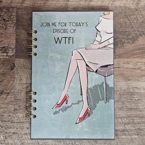 May include: A spiral-bound notebook with a light blue cover featuring a graphic illustration of a person sitting on a stool, wearing a light-colored dress and red high-heeled shoes. The text on the cover reads: JOIN ME FOR TODAY'S EPISODE OF WTF!