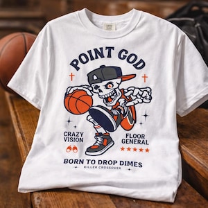 Puede incluir: Camiseta blanca con un gráfico de baloncesto. El diseño presenta un esqueleto con gorra y zapatillas, botando un balón de baloncesto. El texto incluye "POINT GOD", "CRAZY VISION" y "BORN TO DROP DIMES". Un balón de baloncesto y una bolsa negra en el fondo.