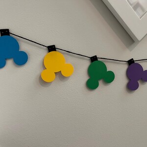 Puede incluir: Una colorida guirnalda con formas de cabeza de Mickey Mouse en azul, amarillo, verde, morado y naranja. Las formas est&aacute;n ensartadas en un cord&oacute;n negro, perfectas para decoraciones de fiestas o eventos tem&aacute;ticos. La guirnalda cuelga contra una pared blanca.