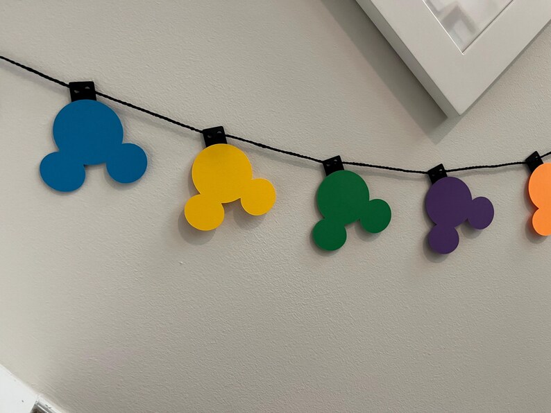 Puede incluir: Una colorida guirnalda con formas de cabeza de Mickey Mouse en azul, amarillo, verde, morado y naranja. Las formas est&aacute;n ensartadas en un cord&oacute;n negro, perfectas para decoraciones de fiestas o eventos tem&aacute;ticos. La guirnalda cuelga contra una pared blanca.