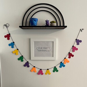 Puede incluir: Una colorida guirnalda de formas de cabeza de Mickey Mouse en rojo, azul, amarillo, verde, morado y naranja cuelga debajo de una impresi&oacute;n enmarcada en blanco que dice "THE PARTY MINIMALIST". Una estanter&iacute;a negra exhibe una maceta azul, una figura morada y una peque&ntilde;a planta.