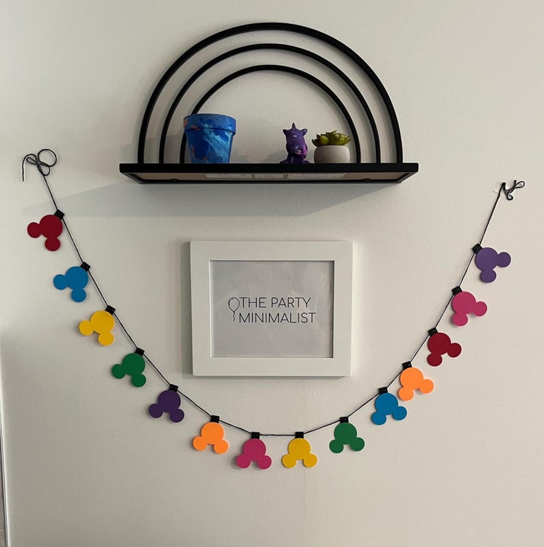 Puede incluir: Una colorida guirnalda de formas de cabeza de Mickey Mouse en rojo, azul, amarillo, verde, morado y naranja cuelga debajo de una impresi&oacute;n enmarcada en blanco que dice "THE PARTY MINIMALIST". Una estanter&iacute;a negra exhibe una maceta azul, una figura morada y una peque&ntilde;a planta.