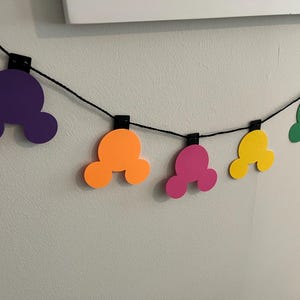Puede incluir: Una colorida guirnalda con formas de cabeza de Mickey Mouse en morado, naranja, rosa, amarillo, verde y azul. Las formas est&aacute;n ensartadas en un cord&oacute;n negro, sujetas con clips negros, creando una decoraci&oacute;n festiva.
