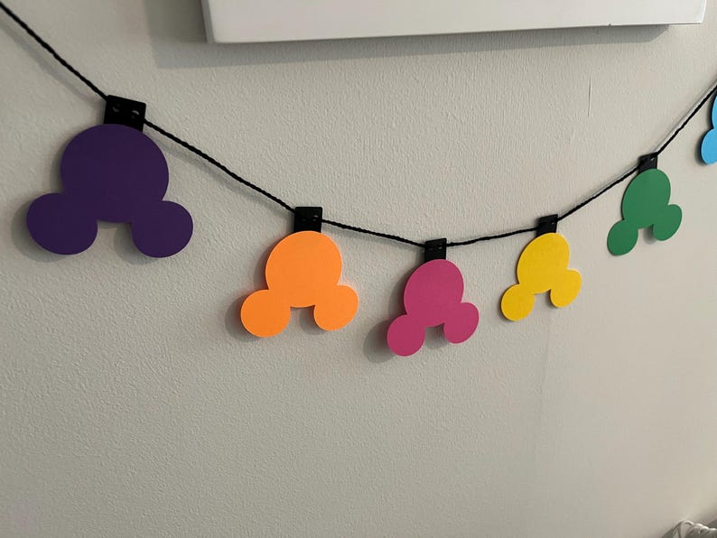Puede incluir: Una colorida guirnalda con formas de cabeza de Mickey Mouse en morado, naranja, rosa, amarillo, verde y azul. Las formas est&aacute;n ensartadas en un cord&oacute;n negro, sujetas con clips negros, creando una decoraci&oacute;n festiva.