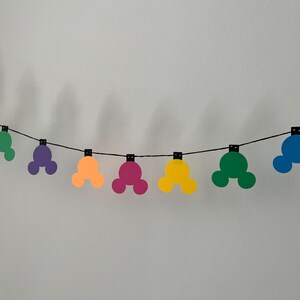 Puede incluir: Una colorida guirnalda con formas de cabeza de Mickey Mouse en amarillo, verde, morado, naranja, rosa, azul y naranja. Las formas est&aacute;n ensartadas a lo largo de una cuerda negra, creando una decoraci&oacute;n festiva. La guirnalda est&aacute; contra una pared blanca lisa.
