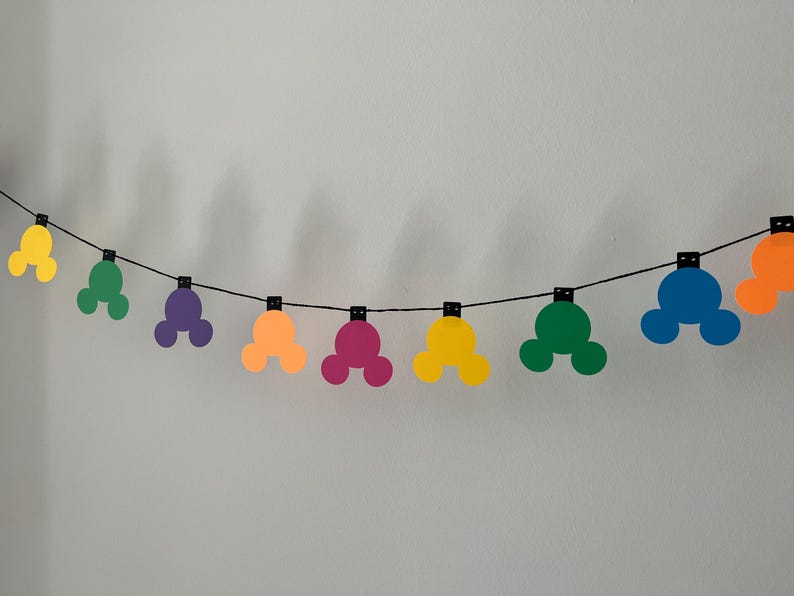 Puede incluir: Una colorida guirnalda con formas de cabeza de Mickey Mouse en amarillo, verde, morado, naranja, rosa, azul y naranja. Las formas est&aacute;n ensartadas a lo largo de una cuerda negra, creando una decoraci&oacute;n festiva. La guirnalda est&aacute; contra una pared blanca lisa.