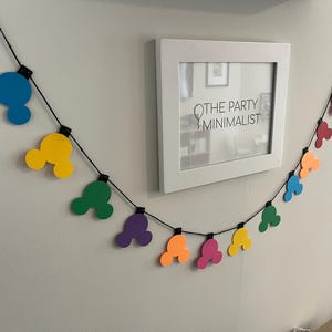 Puede incluir: Una colorida guirnalda de formas de cabeza de Mickey Mouse en rojo, azul, amarillo, verde, morado, naranja y rosa cuelga de una pared. Tambi&eacute;n hay una impresi&oacute;n enmarcada en blanco con el texto "THE PARTY MINIMALIST".