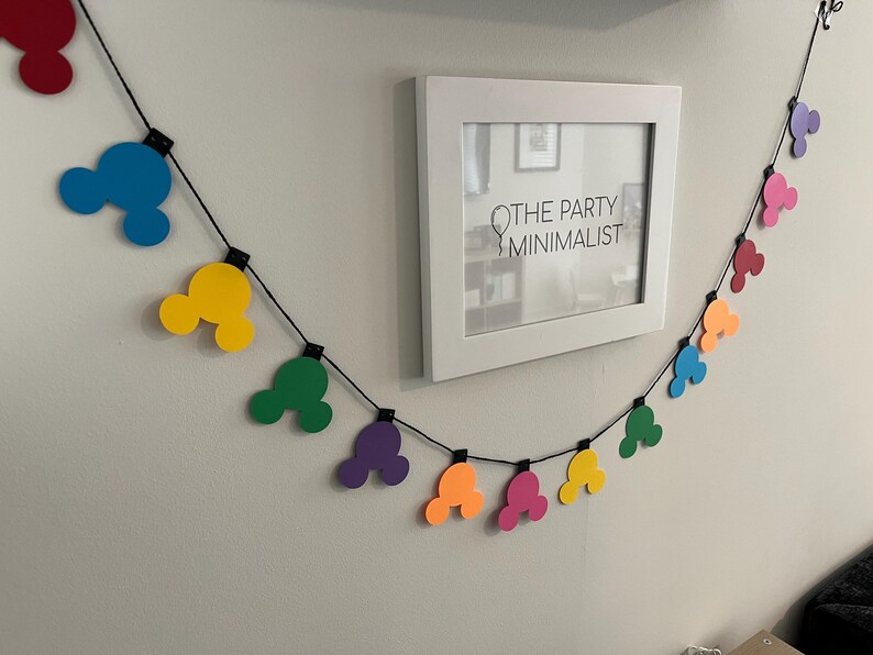 Puede incluir: Una colorida guirnalda de formas de cabeza de Mickey Mouse en rojo, azul, amarillo, verde, morado, naranja y rosa cuelga de una pared. Tambi&eacute;n hay una impresi&oacute;n enmarcada en blanco con el texto "THE PARTY MINIMALIST".
