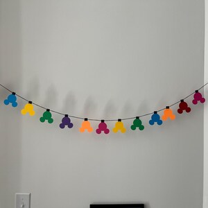 Puede incluir: Una guirnalda colorida de formas de cabeza de Mickey Mouse en rojo, azul, amarillo, verde, morado, naranja y rosa. Las formas est&aacute;n ensartadas en un alambre negro, creando una decoraci&oacute;n festiva. La guirnalda cuelga contra una pared blanca.