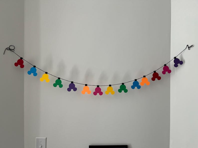 Puede incluir: Una guirnalda colorida de formas de cabeza de Mickey Mouse en rojo, azul, amarillo, verde, morado, naranja y rosa. Las formas est&aacute;n ensartadas en un alambre negro, creando una decoraci&oacute;n festiva. La guirnalda cuelga contra una pared blanca.